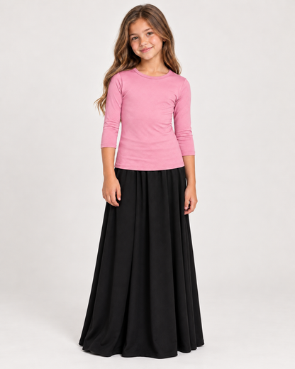 A young girl modeling a three-quarter sleeve pink top and a long black skirt against a plain light gray studio background.
