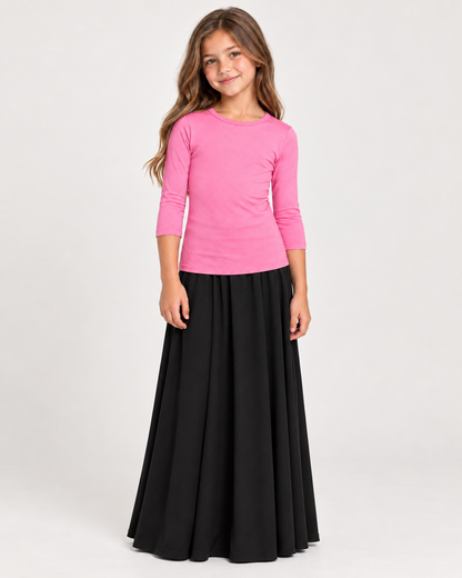 A young girl modeling a bright pink three-quarter sleeve top paired with a long black skirt, standing against a plain light gray studio background.