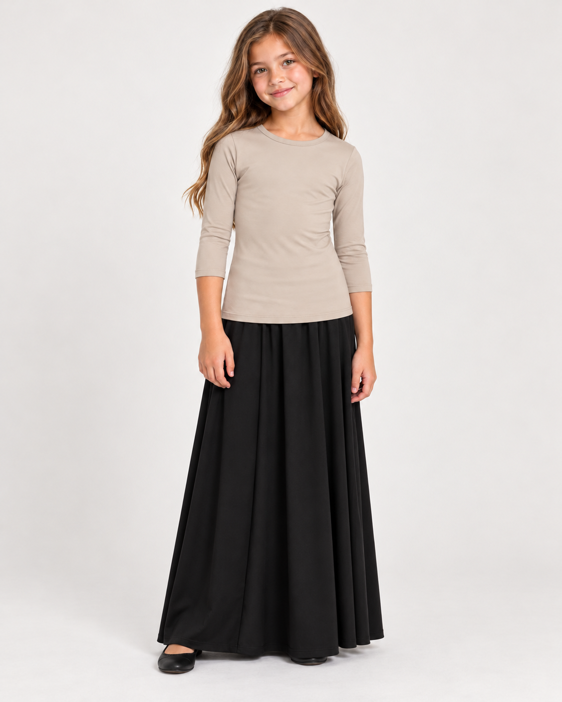 A young girl modeling a three-quarter sleeve beige top and a long black skirt, standing against a plain light gray studio background.