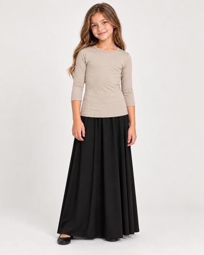 A young girl modeling a three-quarter sleeve beige top and a long black skirt, standing against a plain light gray studio background.