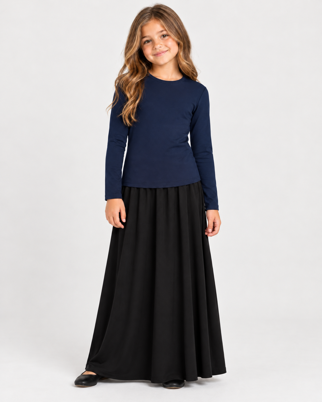 A young girl with long light brown hair stands against a light gray studio background wearing a navy blue long-sleeve top and a long black skirt.