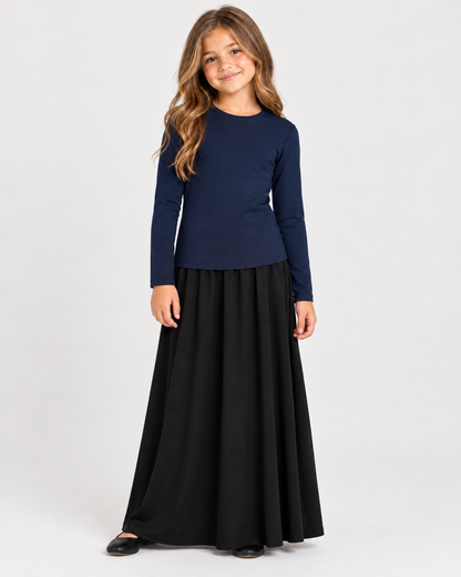A young girl with long light brown hair stands against a light gray studio background wearing a navy blue long-sleeve top and a long black skirt.