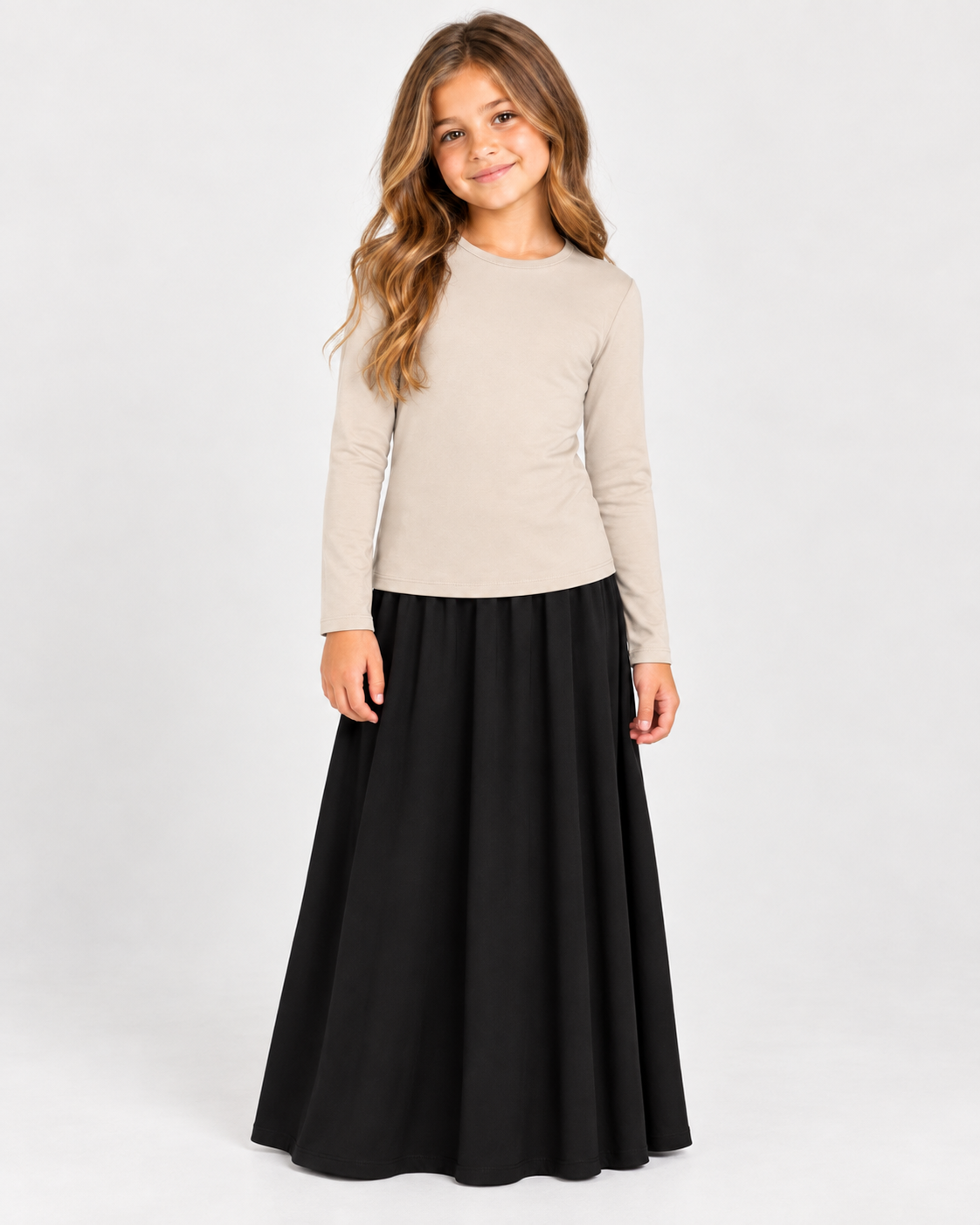 A young girl with long wavy brown hair stands against a light gray studio background, wearing a beige long-sleeve crewneck top and a long black skirt.