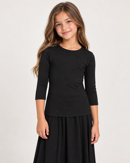 A young girl with long light brown hair wearing a black three-quarter sleeve top and a black skirt, standing against a plain light gray studio background.