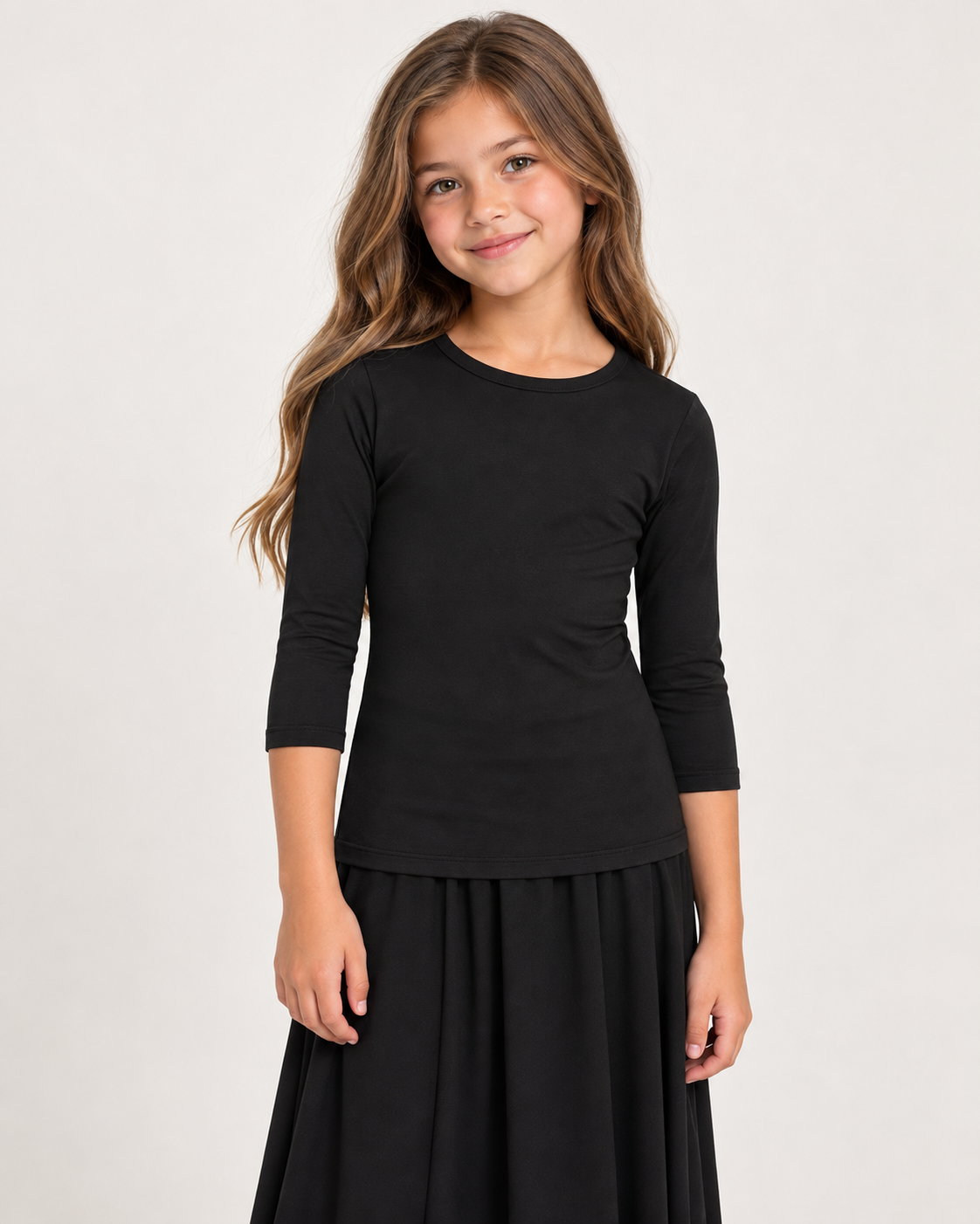 A young girl with long light brown hair wearing a black three-quarter sleeve top and a black skirt, standing against a plain light gray studio background.