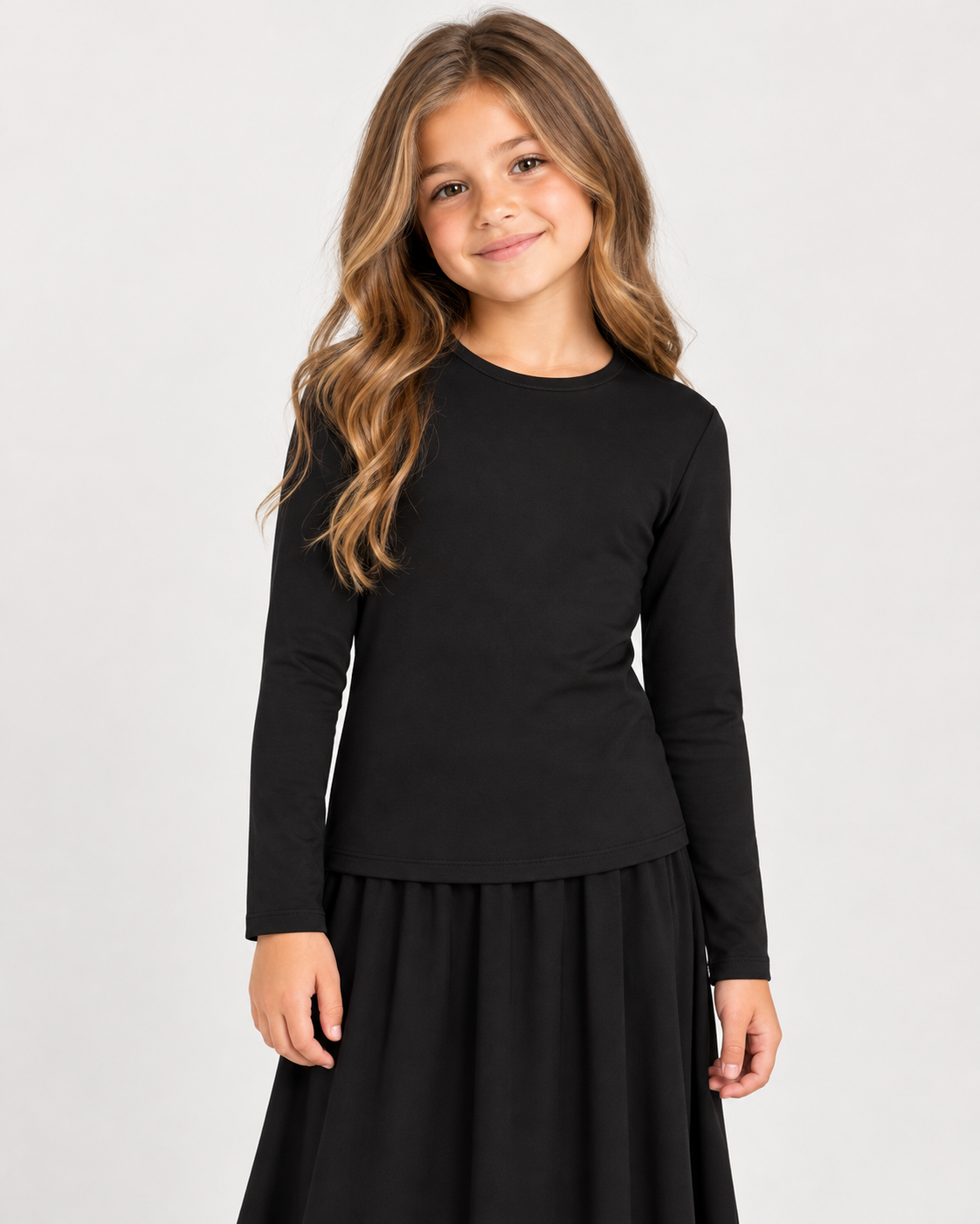 A young girl with long wavy brown hair wearing a black long-sleeve crewneck top and a black skirt, standing against a light gray studio background.