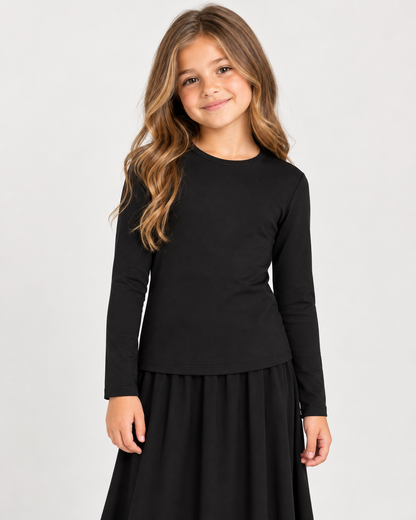 A young girl with long wavy brown hair wearing a black long-sleeve crewneck top and a black skirt, standing against a light gray studio background.