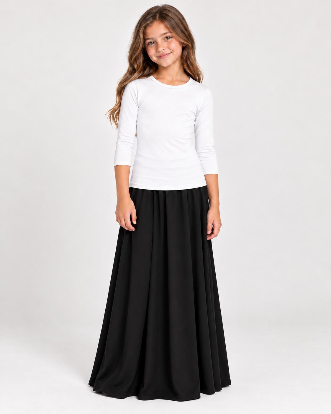 A young girl modeling a white long-sleeve crewneck top and a long black pleated skirt, standing against a light gray studio background.