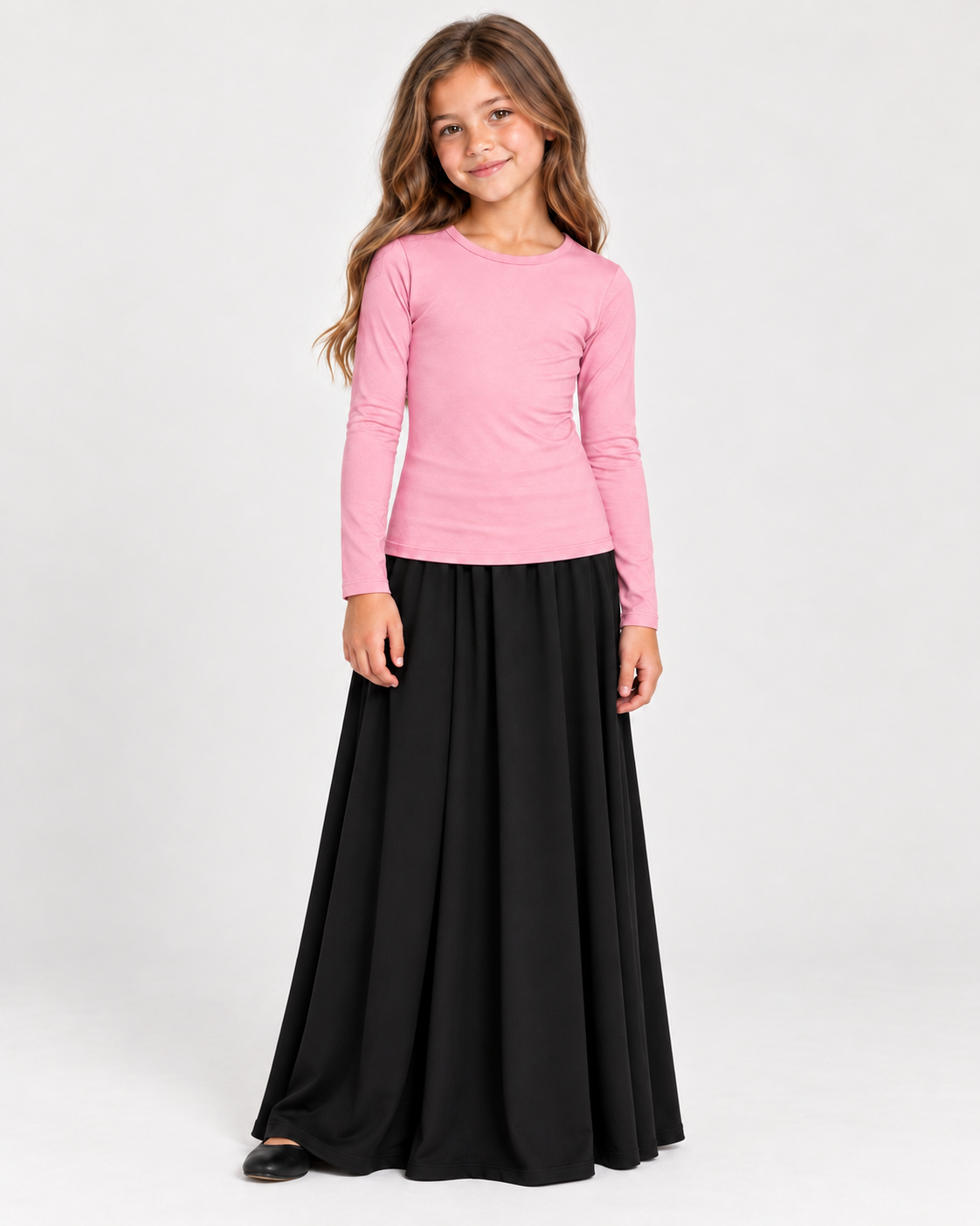 A young girl modeling a pink long-sleeve crewneck top and a long black skirt against a plain light gray studio background.