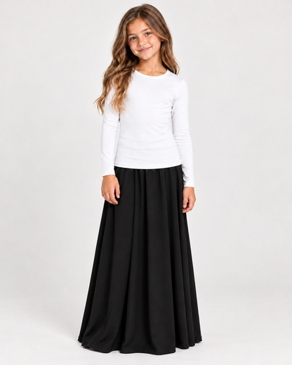 A young girl with long wavy brown hair stands against a light gray studio background, wearing a fitted white long-sleeve top and a long black pleated skirt.