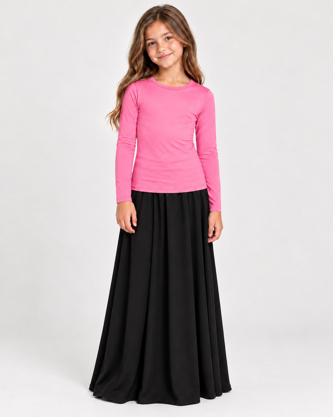 A young girl modeling a bright pink long-sleeve crewneck top paired with a long black skirt, standing against a plain light gray studio background.