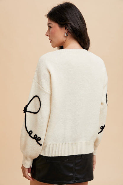 Ivory sweater with black ribbon bow and embroidered trim by ModestAndDevoted, showcasing elegant knit design and detailed accents.