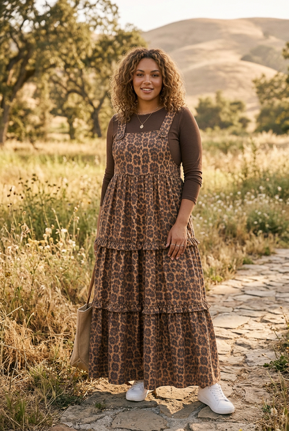 A woman stands outdoors on a stone path in a grassy field with hills in the background, wearing a brown leopard-print tiered maxi dress over a dark brown long-sleeve top and white sneakers.