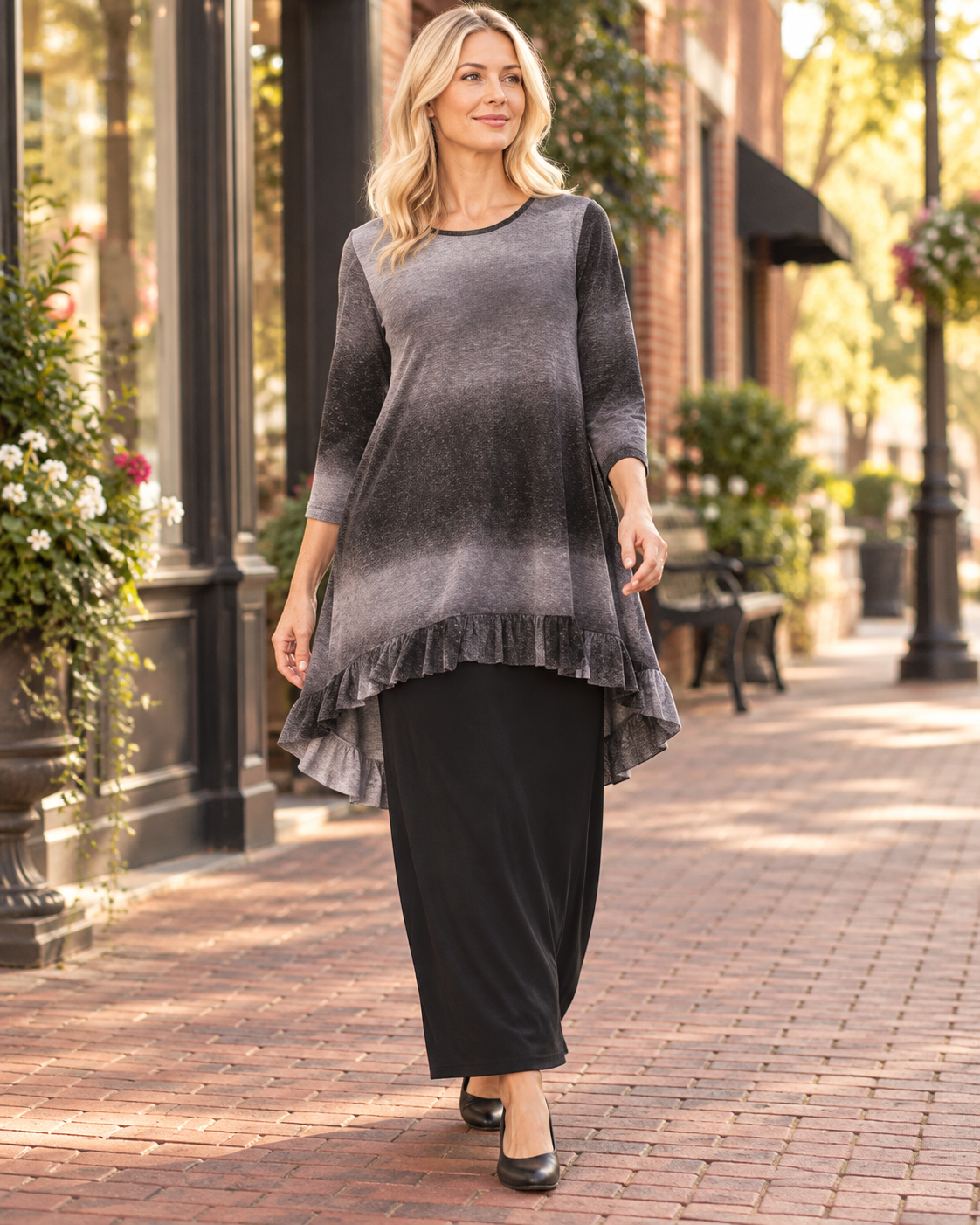 A woman walking on a brick sidewalk wearing a dark gray ombre tunic top with a ruffled hem and black wide-leg pants.